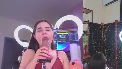 Media: Video of a young woman with long dark hair, wearing a red top, using a glass dildo in a brightly lit room with a large, circular mirror.