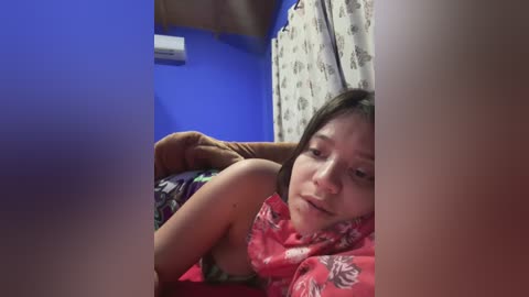 Media: Video of a young Asian woman with dark hair lying on a bed, partially covered by a floral-patterned red blanket, in a blue-walled room with a white curtain and brown cushion.