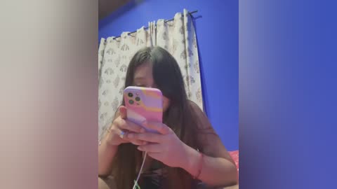 Media: Video of a young woman with long black hair, wearing a striped phone case, taking a selfie in a blue-walled room with white patterned curtains.