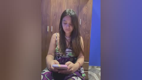 Media: A video of a young Asian girl with long dark hair, wearing a colorful patterned dress, sitting on a bed with wooden paneling and blue walls, engrossed in a tablet.