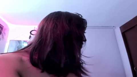 Media: Video of a woman with shoulder-length, dark brown hair, seen from behind in a dimly lit room with white walls, a partially visible window, and a closed white door.