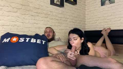 Media: Video of a nude, muscular man with a beard and a tattooed woman with long black hair performing oral sex on him in a bedroom with white brick walls and a dark headboard.