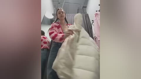 Media: Video of a young woman in a pink, striped cardigan and high-waisted jeans, holding a beige, puffy jacket in a clothing store with racks of clothes in the background.