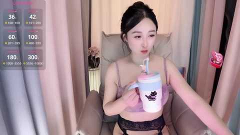 Media: A young Asian woman with fair skin, black hair tied back, wearing a pink bra and black lace panties, sipping from a pink cup in a cozy room with beige curtains and a pink flower.