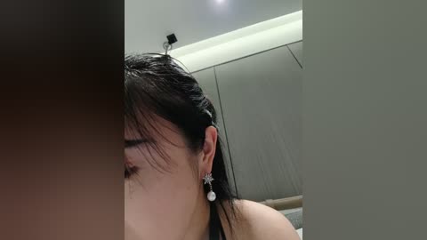 Media: A video of a woman with wet, dark hair, wearing star-shaped earrings, partially obscured by another person's arm, in a modern, dimly lit bedroom with white walls and a large wardrobe.