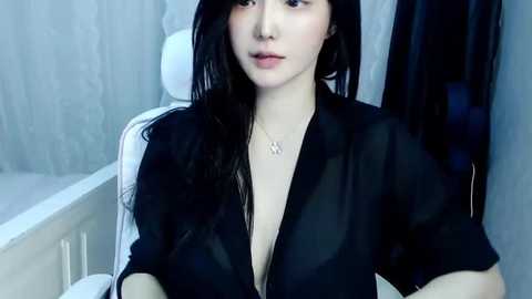 Media: Video of a young Asian woman with long black hair, wearing a black blouse, sitting on a white bed. The background features light blue curtains and a white lamp.