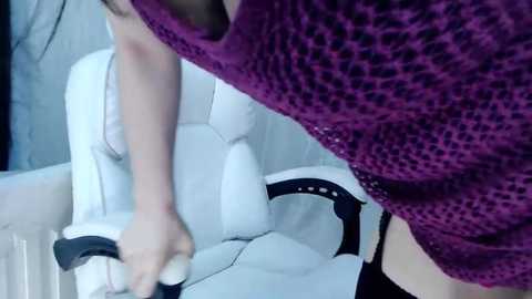 Media: A video captures a person in a purple knit sweater, leaning over a white leather chair, with a black dress and white shoes visible.