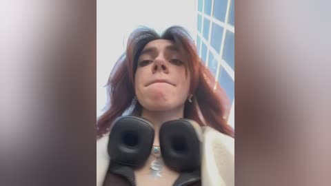 Media: Video of a person with shoulder-length auburn hair, wearing black over-the-ear headphones and a light-colored jacket, taking a selfie with a neutral expression, in a room with blue-framed windows.