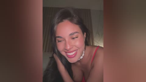 Media: A close-up video of a smiling, young woman with long, dark hair, wearing a red spaghetti-strap top and a white choker necklace.