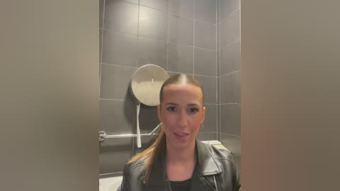 Media: A video of a young woman with light skin and straight brown hair tied back, wearing a black leather jacket and a black top, standing in a tiled bathroom with a metallic soap dispenser mounted on the wall.