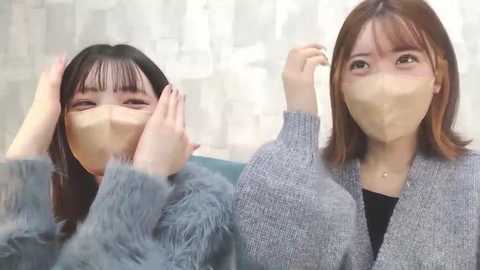 Media: Video of two East Asian women with face masks, one with long black hair, the other with shoulder-length brown hair, both smiling and touching their hair. They wear casual grey sweaters, in a softly lit room with semi-transparent curtains.