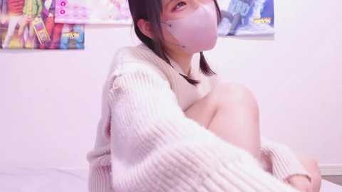 Media: Video of an East Asian woman with short dark hair, wearing a beige sweater, face mask, and no pants, sitting in a brightly lit room with colorful posters in the background.