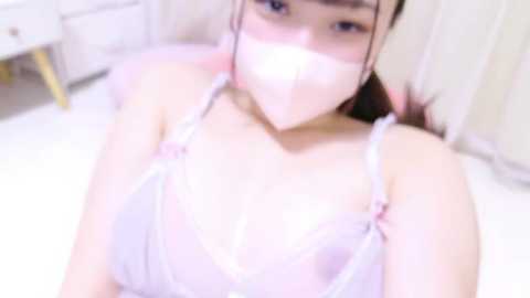 Media: A video of an Asian woman with a fair complexion, wearing a pink, sheer bra, and a face mask, lying on a bed in a bright, minimalistic room.