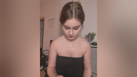 Media: Video of a young Caucasian woman with fair skin and brown hair in a bun, wearing a strapless black dress, standing in a cluttered, dimly-lit room with a white door and a laundry basket.