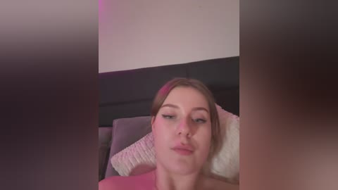 Media: A video of a young woman with fair skin and light brown hair, wearing pink lipstick, lying on a bed with a gray headboard, pillows, and a pink blanket.