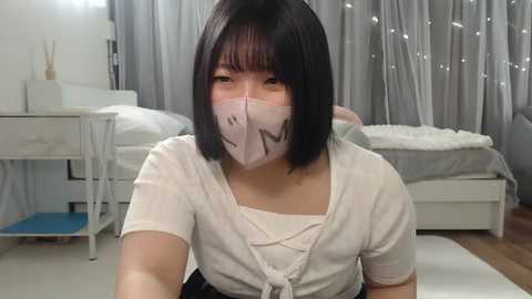 Media: Video of an East Asian woman with straight black hair, wearing a white mask, white top, and gray pants, sitting on a light-colored rug in a modern, minimally decorated bedroom with white furniture, gray curtains, and a bed.