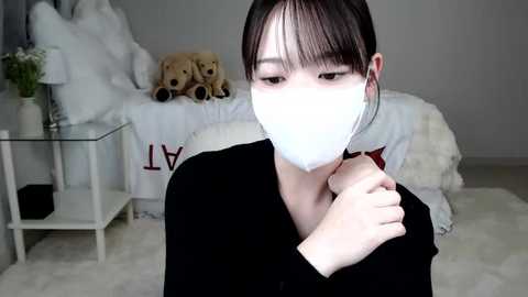 Media: Video of an East Asian woman with fair skin, straight black hair, wearing a white face mask and black top, holding a red mask, sitting on a fluffy white rug in a minimalist bedroom with stuffed toys and a white bed.