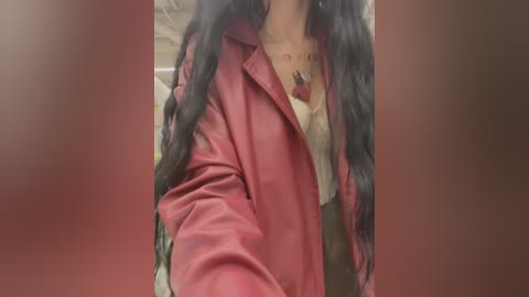 Media: A video of a person with long, wavy black hair, wearing a red leather jacket over a white blouse with a visible tattoo on their chest. The background is blurred, with a beige and red color palette.