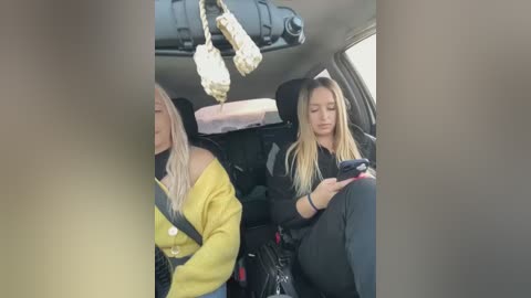 Media: Video of a blonde woman in a yellow jacket and another woman in black clothing, both sitting in a car, with a bag hanging from the ceiling.