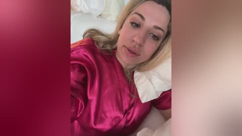 Media: Video of a fair-skinned blonde woman with straight hair, wearing a shiny pink satin robe, lying on a bed with white pillows and a red background.