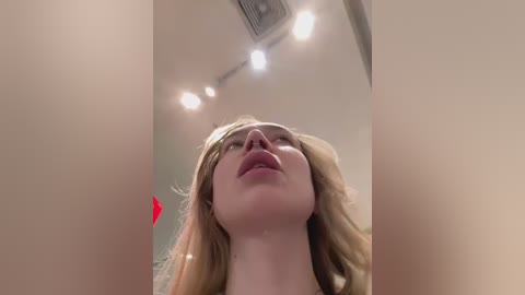 Media: Video of a blonde woman with fair skin, looking up with parted lips, surrounded by white ceiling lights, slightly blurry.