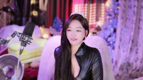 Media: Video of a young Asian woman with long dark hair, wearing a black leather jacket, standing in a dimly lit room with soft purple lighting, surrounded by soft toys and a microphone.