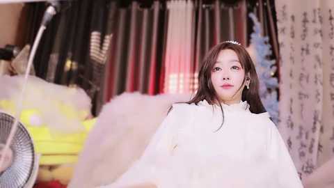 Media: Video of an East Asian woman with long, straight brown hair, wearing a white robe, standing in a softly lit room with curtains, a fan, and a yellow object in the background.