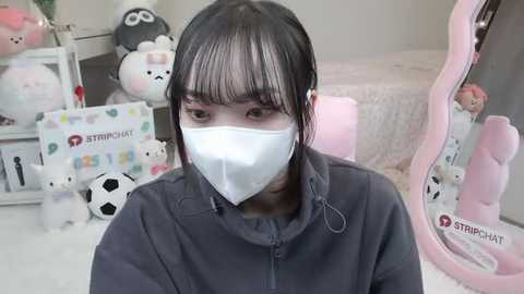 Media: Video of an Asian woman with straight black hair, wearing a white face mask, a dark hoodie, and sitting in a pastel-themed room filled with plush toys and a pink bean bag.