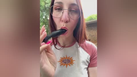 Media: Video of a young woman with fair skin, glasses, and long red hair, wearing a white and pink t-shirt with a sun graphic. She is outdoors, sucking on a black dildo, surrounded by lush greenery.