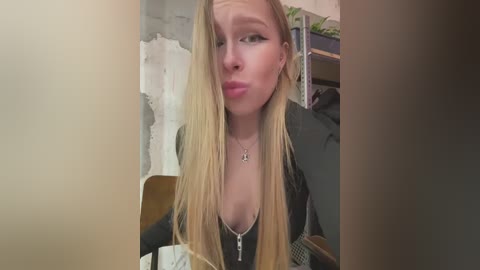 Media: A video of a blonde woman with long hair, wearing a black top and a cross necklace, pouting while indoors with a brick wall and shelves in the background.