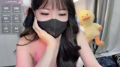 Media: Video of an Asian woman with long black hair, wearing a black face mask, pink lace bra, and white furry headband, holding her chin, surrounded by stuffed toys and a fitness tracker display in a cozy room.