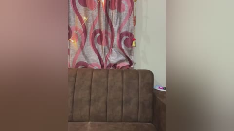 Media: Video of a brown, tufted couch with vertical ridges in a living room. Behind it, a curtain with a red and white floral pattern and yellow tassels hangs against a cream-colored wall.