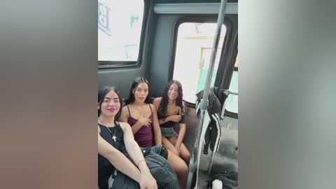 Media: Video of three young women sitting in a public bus. One is Asian with straight black hair, wearing a black top. The other two are Latina with long hair, one in a maroon top, the other in a black top.