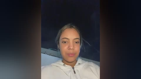 Media: Video of a young woman with medium brown skin, light brown hair pulled back, wearing a white hoodie, against a dark background.