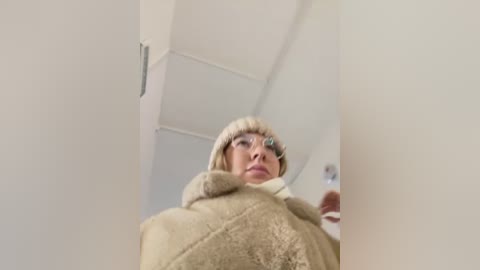 Media: A low-angle video of a blonde woman with a fur-trimmed beige coat, wearing glasses and a fur hat, standing in a minimalist, white-walled room.