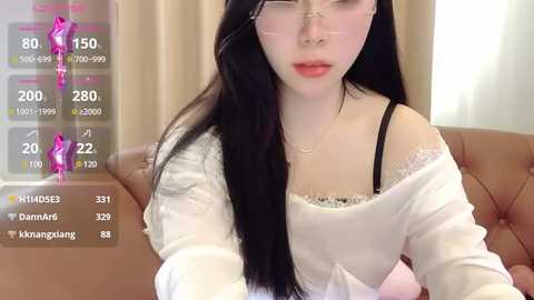 Media: Video of a young Asian woman with long black hair, fair skin, and glasses, wearing a white off-shoulder top, sitting on a brown couch. Digital stats overlay show heart rate, temperature, and activity levels.