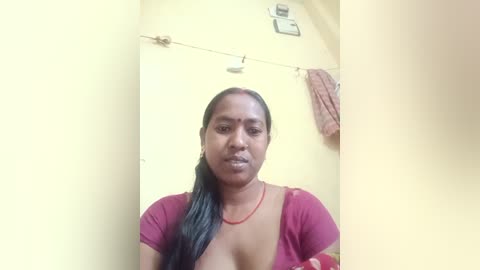 Media: A video of an Indian woman with medium brown skin, wearing a maroon blouse and a red string necklace, standing in a yellow-walled room with a clothesline and towel hanging above.