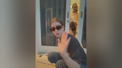 Media: Video of a woman with blonde hair and dark sunglasses, crouching in a doorway, gesturing, and a man in a gray shirt standing behind her in a modern living room.