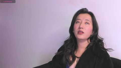 Media: Video of a woman with long black hair and fair skin, wearing a black coat, looking introspective against a plain white background.