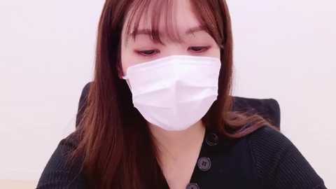 Media: Video of a young East Asian woman with long, straight brown hair, wearing a white surgical mask, black cardigan, and a pale background.