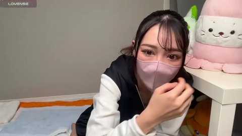 Media: Video of an Asian woman with long black hair, wearing a pink mask, a black vest, and a white shirt, sitting on a bed, leaning against a white table with plush toys.