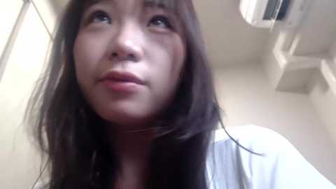 Media: A close-up video of a young East Asian woman with long, straight black hair, light skin, and a neutral expression. She's indoors, wearing a white top, with a white ceiling and air conditioning unit visible in the background.