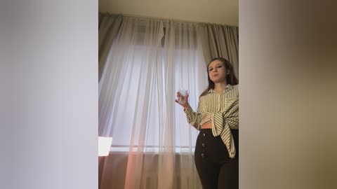 Media: A video of a woman with long brown hair, wearing a striped blouse and high-waisted black pants, gesturing with her hands, standing in front of a window with sheer white curtains.