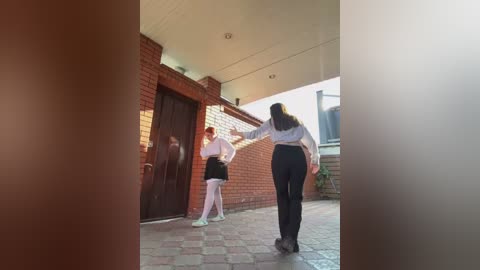 Media: Video of two women, one in a white blouse and black skirt, the other in a white blouse and black pants, walking away from a brick building with a large wooden door.