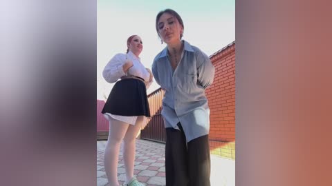 Media: Video of two women in casual attire, one in a white blouse and black skirt, the other in a light blue shirt and black pants, standing on a rooftop with brick walls and a tiled floor.