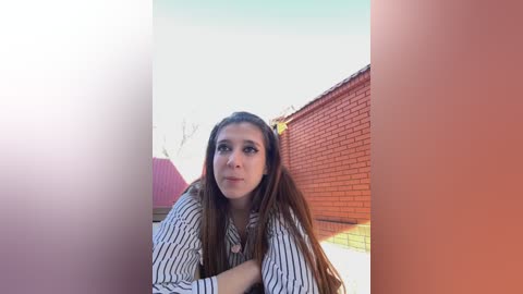 Media: Video of a young woman with long brown hair, wearing a striped shirt, sitting against a red brick wall and a pink roof, under a clear blue sky.