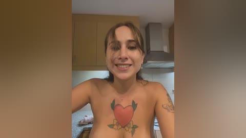 Media: Video of a smiling, topless woman with light brown skin and medium-length brown hair. She has a tattoo of a heart with leaves on her chest. Background shows a kitchen with wooden cabinets and a stove.