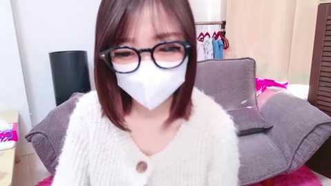 Media: Video of an Asian woman with straight, shoulder-length brown hair, wearing black-framed glasses, a white mask, and a beige knitted cardigan. She sits on a gray couch in a room with a wooden cabinet, pink bedding, and a black floor lamp.