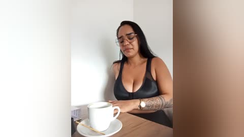 Media: Video of a middle-aged woman with long black hair, glasses, and tattoos, wearing a black sleeveless top, holding a white coffee cup at a wooden table.