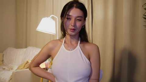 Media: Video of an Asian woman with long black hair, wearing a white halter top, standing in a dimly-lit bedroom with beige curtains and a lamp.